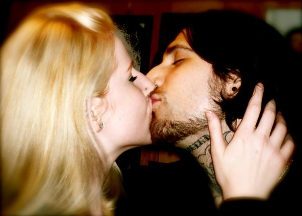 Alexis Smith, with flowing blonde hair, and Banksy with black hair and tattoos on his neck, face each other and kiss, Alexis has a hand on Banksy's neck