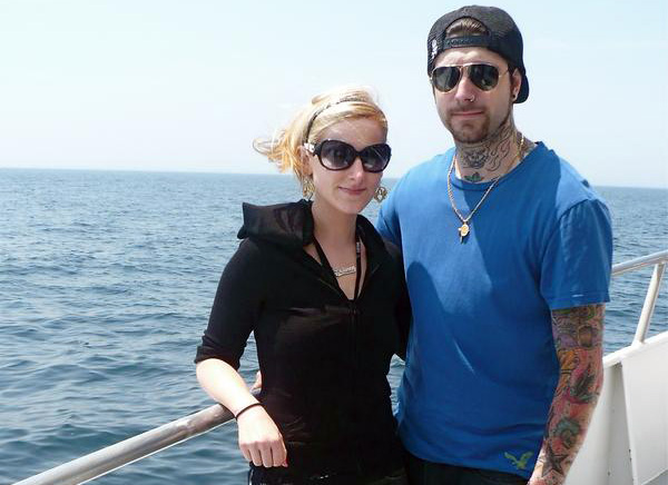 Two people, Alexis Fallon Smith in a black top, and Banksy in a blue t-shirt, stand together at the bow of a boat. They face the camera with an expanse of ocean behind them.