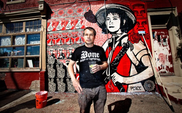 Shepard Fairey standing in front of a just-wheat-pasted mural on the side of the Rock Cat Cafe building in Philadelphia. The iconic black, white, and red poster imagery features his familiar imagery and a very large image of an Asian woman with a hat and a machine gun with a flower blooming out of the bun barrel, a al Bernie Boston
